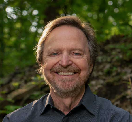 A middle-aged man with light brown hair and a beard smiles outdoors, wearing a dark shirt. The background is lush and green with trees and foliage, suggesting a natural, wooded setting.